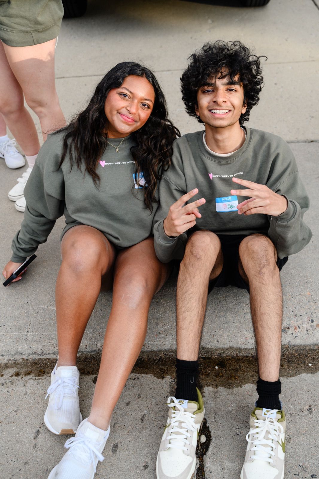 Two teens sitting on a curb, one making a peace sign