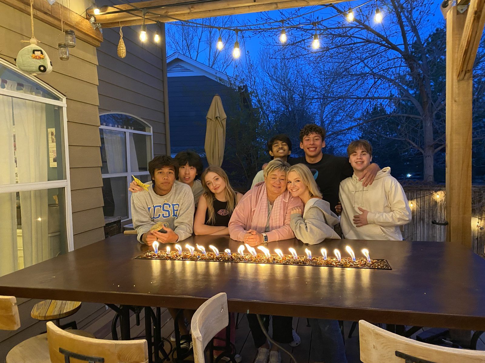 Amy with teens at her backyard table, a line of flames down the middle, string lights overhead